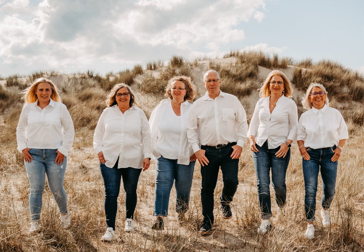Eine Gruppe von Menschen läuft nebeneinander und schaut in die Kamera. Im Hintergrund Himmel, Wolken und eine Dünenlandschaft.